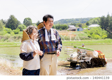 一對老年夫婦面帶微笑地站在一台插秧機前,背景是日本鄉村景觀。 一對老年夫婦面帶微笑地站在一台插秧機前,背景是日本鄉村景觀。 140441139