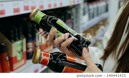 Customer comparing two bottles of soy sauce, reading labels and making purchasing decision in grocery store, showcasing consumer behavior and product selection process 140441415