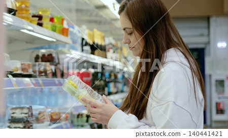 Young woman carefully examining the label of a food product, deciding what to buy in the refrigerated section of a supermarket Young woman carefully examining the label of a food product, deciding what to buy in the refrigerated section of a supermarket 140441421