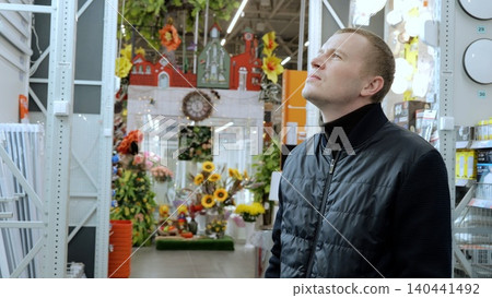 A man in a hardware store, choosing materials for repairs. Man carefully selects lighting for his apartment while standing in a hardware store near the counter and examining products on the shelves A man in a hardware store, choosing materials for repairs. Man carefully selects lighting for his apartment while standing in a hardware store near the counter and examining products on the shelves 140441492