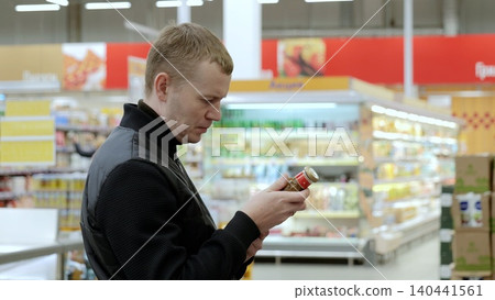 Careful customer reads product label in brightly lit supermarket aisle, making thoughtful healthy choice. A man studies the label on a bottle of sauce.Thoughtful customer carefully reads product label 140441561