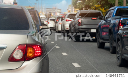 Vehicles are gridlocked in heavy daytime traffic on city asphalt road. A silver car is prominent on the left with dark pickup trucks and sedans forming long rows ahead. Blurred of city background. 140441731