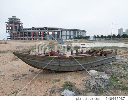 Dilapidated boat left on the shore near a modern construction site under a cloudy sky. 140442987