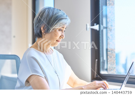 A senior woman in a white shirt is concentrating on her computer work by the window. 140443759