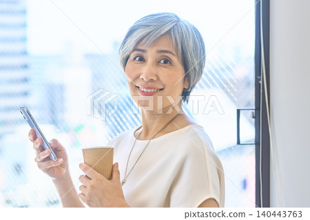 A senior woman smiles by the window, holding her smartphone and a paper cup. 140443763