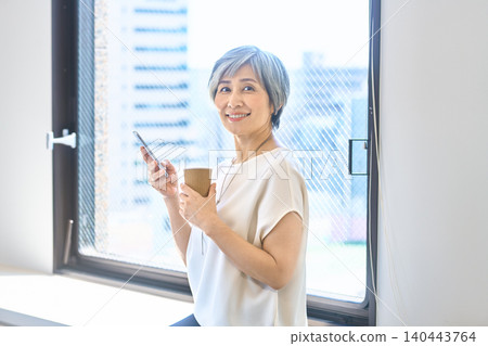 A smiling senior woman holding a smartphone and a paper cup by the window. 140443764