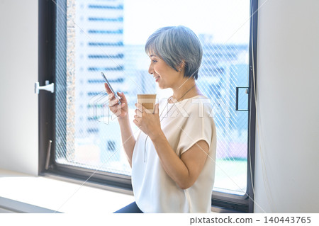 A senior woman sitting by the window, looking at her smartphone and holding a cup of coffee. 140443765