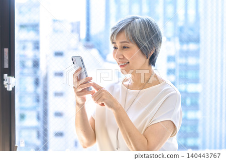 A senior woman with gray hair laughs while operating her smartphone by the window. A senior woman with gray hair laughs while operating her smartphone by the window. 140443767
