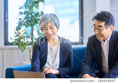 A female consultant and a senior man are seen discussing something while looking at a laptop on a sofa. A female consultant and a senior man are seen discussing something while looking at a laptop on a sofa. 140443841