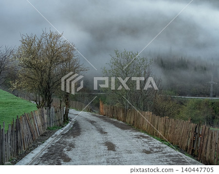 Foggy morning in village. Damp gravel path winds between rustic wooden fences Foggy morning in village. Damp gravel path winds between rustic wooden fences 140447705