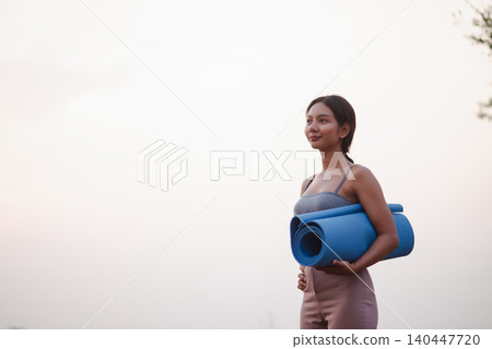 Young woman holding a rolled blue yoga mat near a lake after outdoor practice, representing wellness lifestyle, fitness routine, mindfulness, healthy habits, relaxation, and peaceful nature. Young woman holding a rolled blue yoga mat near a lake after outdoor practice, representing wellness lifestyle, fitness routine, mindfulness, healthy habits, relaxation, and peaceful nature. 140447720