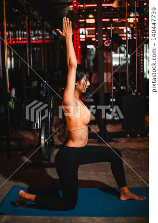 Woman performs a low lunge yoga pose with arms raised on a blue mat in a gym, showing flexibility, balance, strength, stretching, mobility, mindful breathing, and wellness routine. Woman performs a low lunge yoga pose with arms raised on a blue mat in a gym, showing flexibility, balance, strength, stretching, mobility, mindful breathing, and wellness routine. 140447739