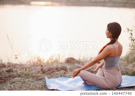 Woman meditating on a blue yoga mat beside a calm lake at sunset, representing outdoor mindfulness, breathing practice, wellness routine, peaceful relaxation, nature, balance, Woman meditating on a blue yoga mat beside a calm lake at sunset, representing outdoor mindfulness, breathing practice, wellness routine, peaceful relaxation, nature, balance, 140447740