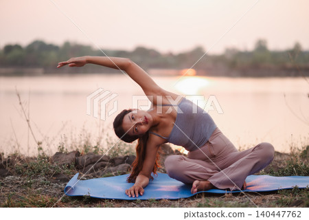 Woman doing a seated side bend on a blue yoga mat beside a lake, emphasizing outdoor wellness, flexibility, core strength, mobility, mindful stretching, calm balance, and healthy lifestyle. 140447762