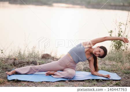 Woman performing a side body yoga stretch on a blue mat by the lake, emphasizing flexibility, core strength, balance, mobility, outdoor fitness, wellness routine, and mindful movement. 140447763