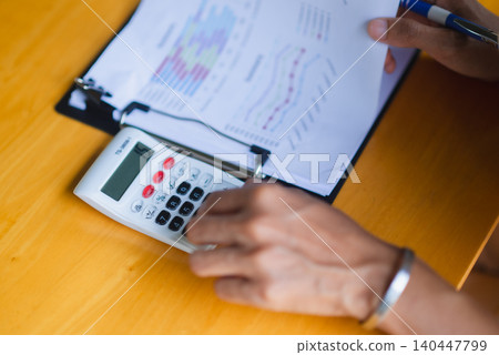 Close-up of hands using a calculator on a financial report with charts, showing budgeting, accounting, tax planning, business analysis, expense tracking, and office paperwork and data review. Close-up of hands using a calculator on a financial report with charts, showing budgeting, accounting, tax planning, business analysis, expense tracking, and office paperwork and data review. 140447799