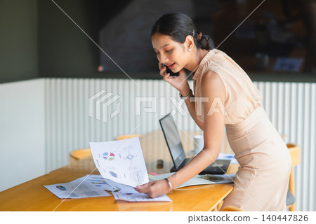 Businesswoman talking on a smartphone while reviewing financial reports on an office desk, showing client communication, budgeting, accounting, decision making, multitasking, and workflow. Businesswoman talking on a smartphone while reviewing financial reports on an office desk, showing client communication, budgeting, accounting, decision making, multitasking, and workflow. 140447826