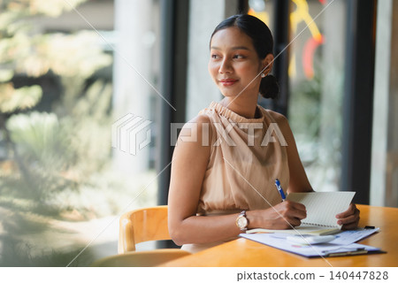 Businesswoman writing notes in a notebook at a modern office desk by large windows, showing planning, productivity, paperwork, financial review, focus, organization, and professional work routine. Businesswoman writing notes in a notebook at a modern office desk by large windows, showing planning, productivity, paperwork, financial review, focus, organization, and professional work routine. 140447828