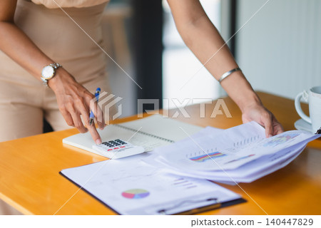 Close-up hands using a calculator while reviewing financial charts on a wooden desk, showing accounting, budgeting, expense tracking, paperwork, audit, business analysis, and tax planning. Close-up hands using a calculator while reviewing financial charts on a wooden desk, showing accounting, budgeting, expense tracking, paperwork, audit, business analysis, and tax planning. 140447829