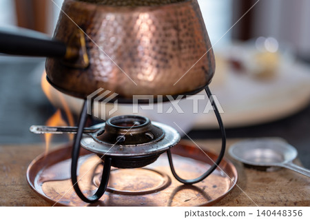 Copper fondue pot on a burner with visible flame heating hot oil, ready for traditional burgundian meat fondue in a cozy restaurant or home kitchen setting Copper fondue pot on a burner with visible flame heating hot oil, ready for traditional burgundian meat fondue in a cozy restaurant or home kitchen setting 140448356