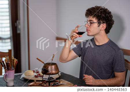 Young man with curly hair and glasses enjoying a relaxing evening at home, drinking red wine and preparing meat in a bourguignonne fondue pot for dinner Young man with curly hair and glasses enjoying a relaxing evening at home, drinking red wine and preparing meat in a bourguignonne fondue pot for dinner 140448363