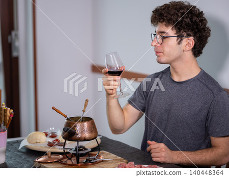 Young man holding a glass of red wine, savoring the aroma before enjoying a traditional meat fondue bourguignonne cooked in boiling oil, with various sauces and fresh bread ready on the dining table Young man holding a glass of red wine, savoring the aroma before enjoying a traditional meat fondue bourguignonne cooked in boiling oil, with various sauces and fresh bread ready on the dining table 140448364