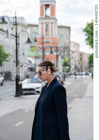 Young woman in dark blazer and sunglasses stands on Pyatnitskaya Street with historic bell tower and urban architecture. City tourism, travel destination, urban lifestyle, street photography Young woman in dark blazer and sunglasses stands on Pyatnitskaya Street with historic bell tower and urban architecture. City tourism, travel destination, urban lifestyle, street photography 140448783