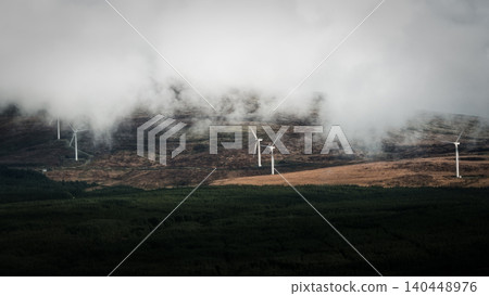 Several wind turbines stand on a misty, rolling hill dotted with brush, with a dark forest in the foreground. 140448976