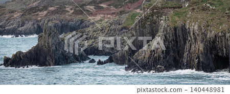 Sharp, vertical rock spires stand in choppy blue water, with vegetation dotting the upper slopes of the surrounding rocky coastline. Sharp, vertical rock spires stand in choppy blue water, with vegetation dotting the upper slopes of the surrounding rocky coastline. 140448981
