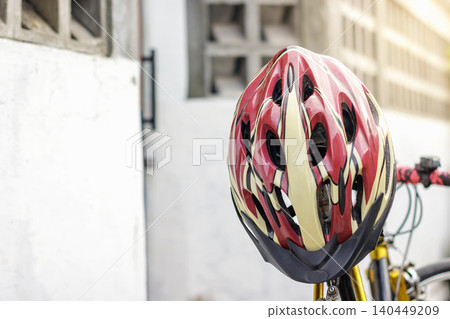 A red and white sport bicycle helmet resting on a saddle with blurred road bike handlebar, positioned in front of a white concrete wall in a bright outdoor alleyway. High Quality Photo 140449209