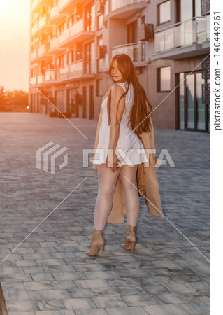 Woman fashion city Young female model posing in a white dress and trench coat on a modern urban street at sunset Woman fashion city Young female model posing in a white dress and trench coat on a modern urban street at sunset 140449261
