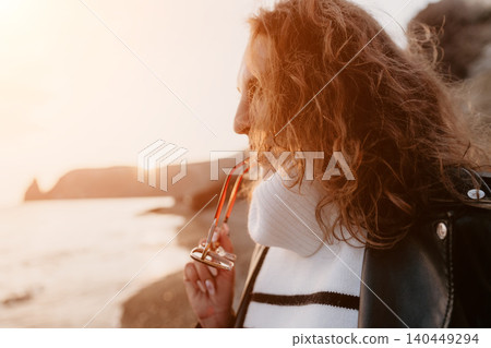 Sunset woman seaside gazing at the ocean horizon during golden hour while holding eyeglasses on a rocky beach Sunset woman seaside gazing at the ocean horizon during golden hour while holding eyeglasses on a rocky beach 140449294