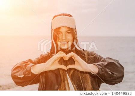 Woman heart gesture young female wearing ushanka makes love sign with hands at sunset sea shore with copy space Woman heart gesture young female wearing ushanka makes love sign with hands at sunset sea shore with copy space 140449883