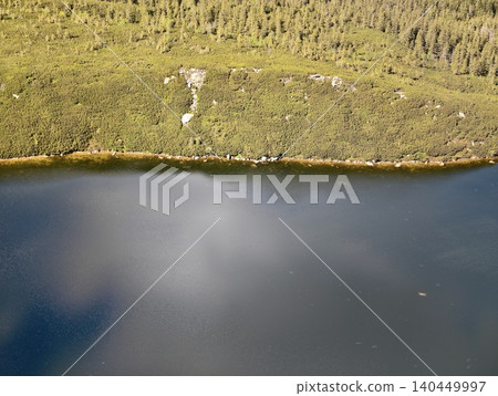 Aerial view of calm lake beside forested hillside and rocky shore 140449997