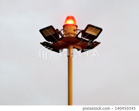 Yellow airport apron light mast with red rotating beacon and floodlights against gray sky 140450345