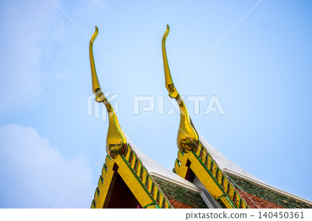 Close up of golden Chofah on traditional Thai temple roof gable with colorful tiles against blue sky background for cultural travel or Asian architectural design concept Close up of golden Chofah on traditional Thai temple roof gable with colorful tiles against blue sky background for cultural travel or Asian architectural design concept 140450361