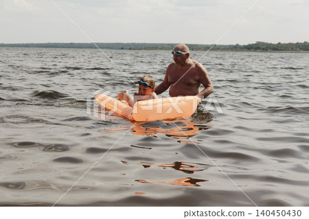 grandfather and grandson enjoy calm summer day. elderly man stands in water holding his child orange inflatable mattress. celebration intergenerational unity, outdoor recreation leisure Family values 140450430