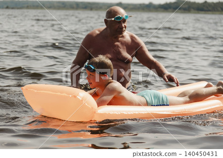 grandfather and grandson enjoy calm summer day. elderly man stands in water holding his child orange inflatable mattress. celebration intergenerational unity, outdoor recreation leisure Family values 140450431
