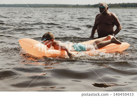 grandfather and grandson enjoy calm summer day. elderly man stands in water holding his child orange inflatable mattress. celebration intergenerational unity, outdoor recreation leisure Family values 140450433