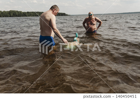 child wearing a blue swimming mask partially submerged in the water father leans over him, gently supporting and assisting. A message about safety, parenting family bonding during outdoor activities 140450451