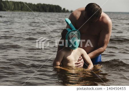 child wearing a blue swimming mask partially submerged in the water father leans over him, gently supporting and assisting. A message about safety, parenting family bonding during outdoor activities child wearing a blue swimming mask partially submerged in the water father leans over him, gently supporting and assisting. A message about safety, parenting family bonding during outdoor activities 140450455