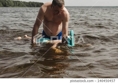 child wearing a blue swimming mask partially submerged in the water father leans over him, gently supporting and assisting. A message about safety, parenting family bonding during outdoor activities child wearing a blue swimming mask partially submerged in the water father leans over him, gently supporting and assisting. A message about safety, parenting family bonding during outdoor activities 140450457
