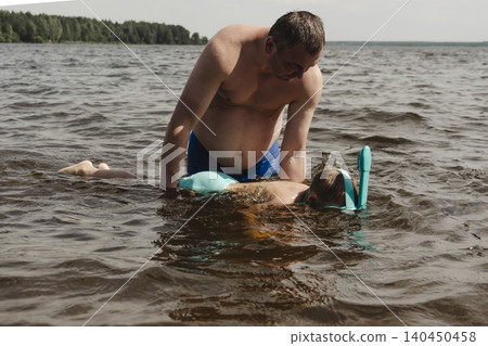 child wearing a blue swimming mask partially submerged in the water father leans over him, gently supporting and assisting. A message about safety, parenting family bonding during outdoor activities child wearing a blue swimming mask partially submerged in the water father leans over him, gently supporting and assisting. A message about safety, parenting family bonding during outdoor activities 140450458