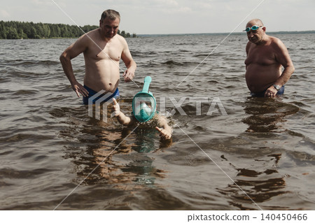 child wearing a blue swimming mask partially submerged in the water father leans over him, gently supporting and assisting. A message about safety, parenting family bonding during outdoor activities 140450466
