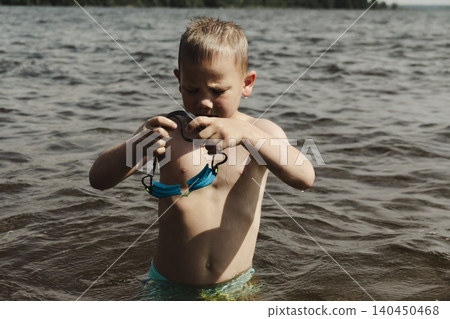 A candid shot of a young boy standing in shallow water at a natural lake or river. He is holding blue swimming goggles in front of his face, preparing to put them on. captures the anticipation 140450468
