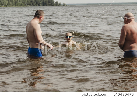 family moment at a natural lake in summer. Two adults, father and grandfather, stand in shallows, carefully watchin child swimming nearby. theme is family vacation, parenting, summer fun tourism. family moment at a natural lake in summer. Two adults, father and grandfather, stand in shallows, carefully watchin child swimming nearby. theme is family vacation, parenting, summer fun tourism. 140450476