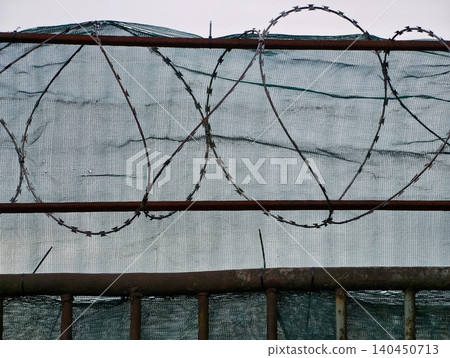 Rusty metal fence topped with coiled barbed wire and green mesh backdrop Rusty metal fence topped with coiled barbed wire and green mesh backdrop 140450713