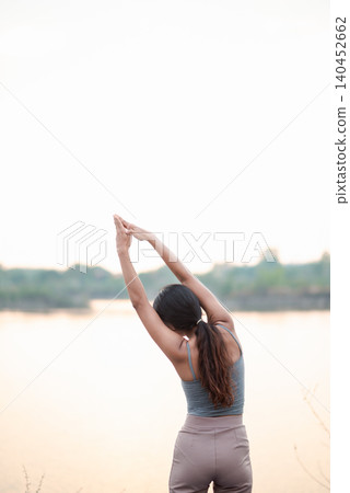 Back view of an Asian woman stretching arms overhead on a blue yoga mat beside a quiet lake, showing outdoor wellness, morning flexibility, mindful breathing, relaxation, calm balance, Back view of an Asian woman stretching arms overhead on a blue yoga mat beside a quiet lake, showing outdoor wellness, morning flexibility, mindful breathing, relaxation, calm balance, 140452662