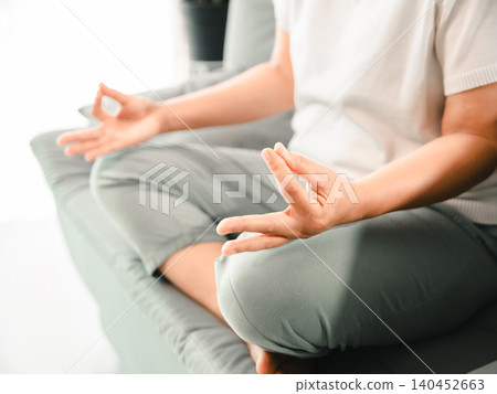 Close-up of Asian woman hands in meditation mudra while sitting cross-legged on a sofa at home, showing mindfulness, calm breathing, stress relief, inner balance, wellness, relaxation, Close-up of Asian woman hands in meditation mudra while sitting cross-legged on a sofa at home, showing mindfulness, calm breathing, stress relief, inner balance, wellness, relaxation, 140452663