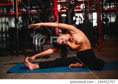 Asian woman stretching in a seated side bend on a yoga mat inside a dark gym, showing flexibility, mobility training, fitness warm up, mindful movement, wellness, strength, and balance. Asian woman stretching in a seated side bend on a yoga mat inside a dark gym, showing flexibility, mobility training, fitness warm up, mindful movement, wellness, strength, and balance. 140452707